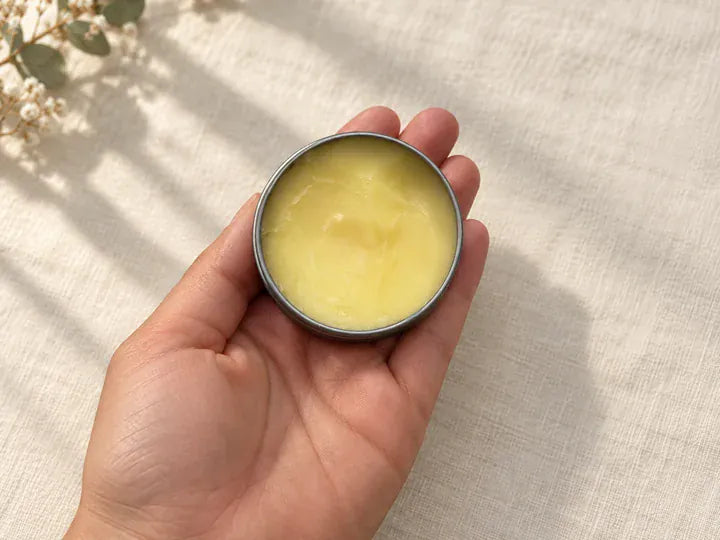 Illustration, not a product photograph: an open balm tin resting in an adult hand