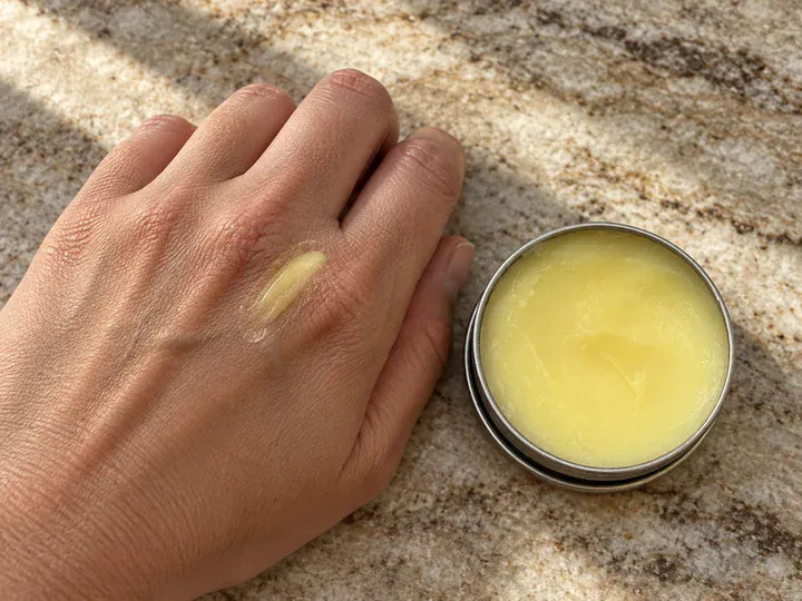 Illustration, not a product photograph: a small amount of balm on a hand beside an open silver tin